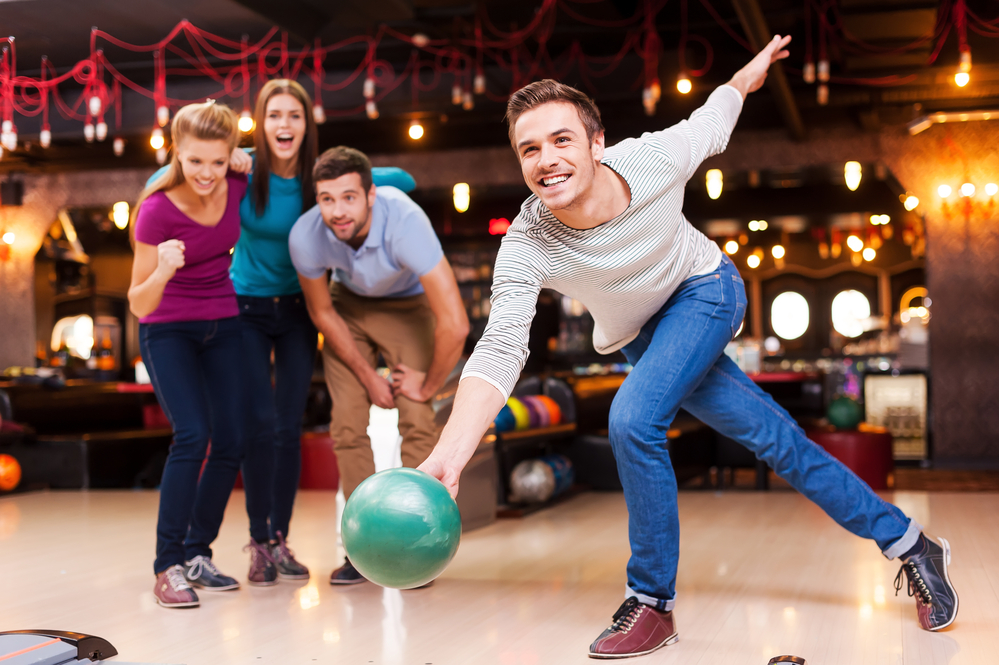 Handsome young man throwing a bowling ball while his friends encourage high scoring.