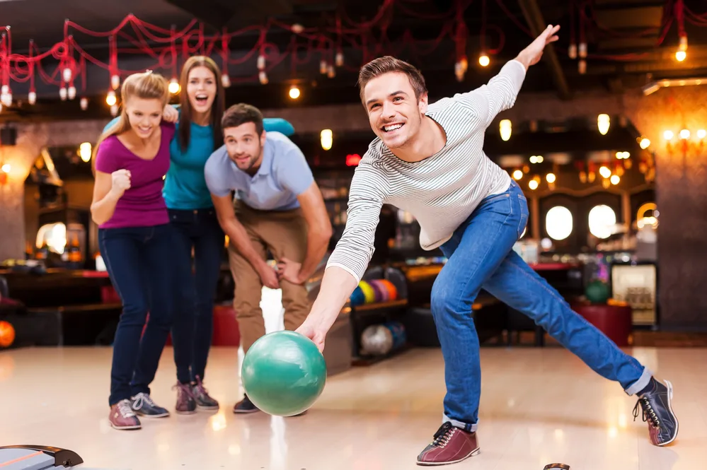 Handsome young man throwing a bowling ball while his friends encourage high scoring.