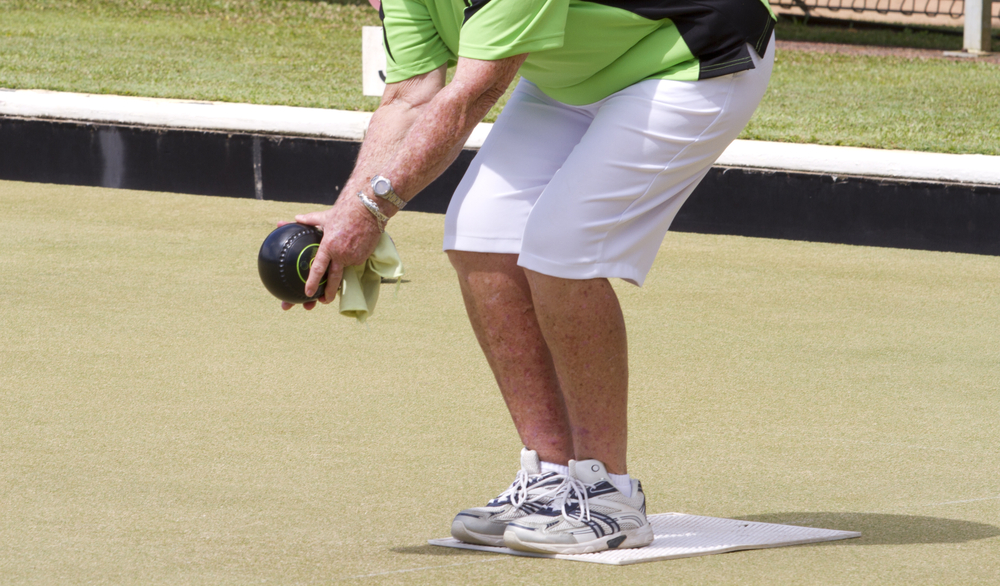 Woman in lime green shirt and white shorts, playing lawn bowling, and outdoor sport, is  holding biased lawned ball on the mat