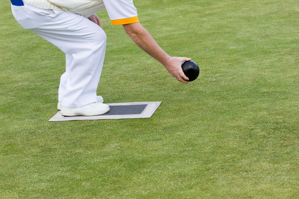 A lawn bowler in all white stands on the mat with the lawn bowl is a right handed bowler