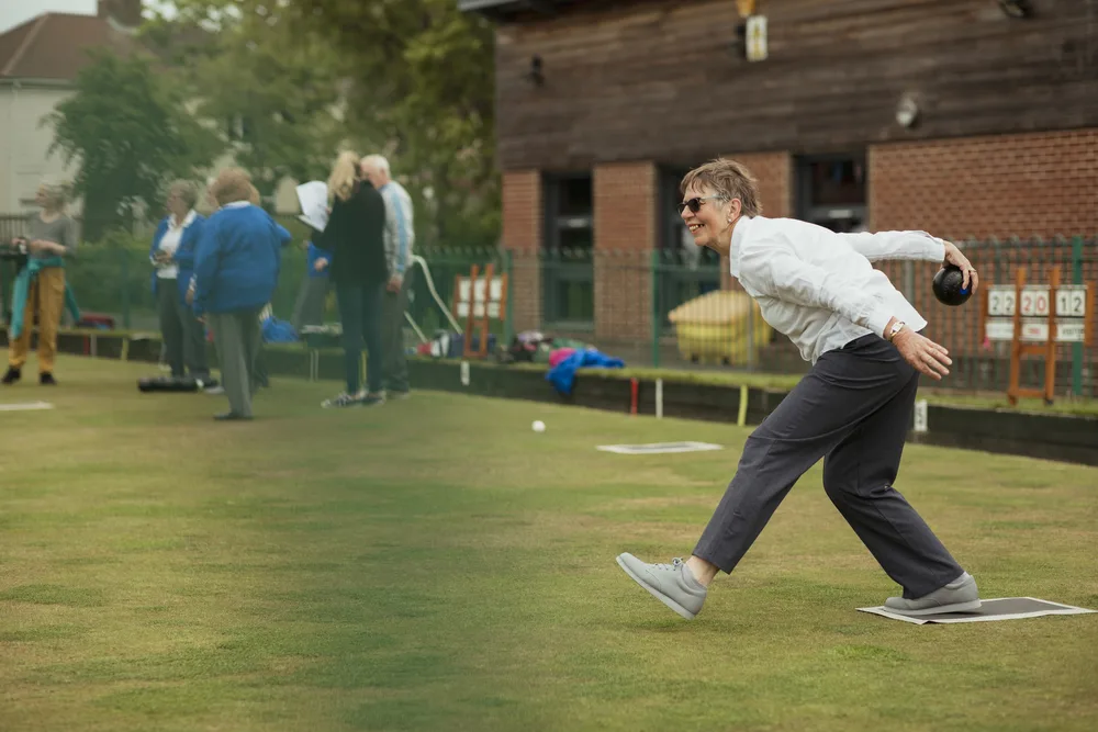 A side view shot of an excited woman in a white shirt taking her shot in a game of lawn bowling.